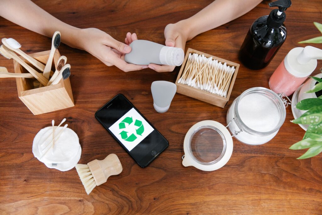 Top view of anonymous person at table with various eco friendly products for skincare and taking tube of cosmetics while preparing for spa procedure near smartphone with recycling symbol at screen