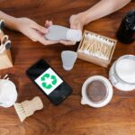 Top view of anonymous person at table with various eco friendly products for skincare and taking tube of cosmetics while preparing for spa procedure near smartphone with recycling symbol at screen
